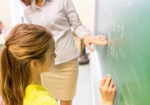 education, elementary school, learning, math and people concept - close up of little schoolgirl with teacher writing on chalk board and solving task at classroom