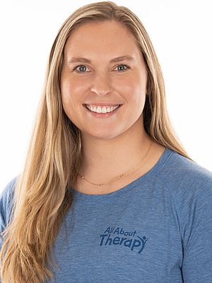Smiling woman in a blue All About Therapy t-shirt with long blonde hair, promoting pediatric occupational therapy services.