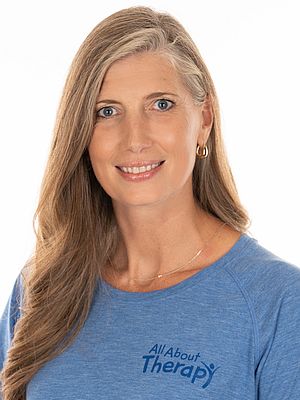 Professional woman with long blonde hair wearing a blue All About Therapy t-shirt, smiling confidently against a white background.