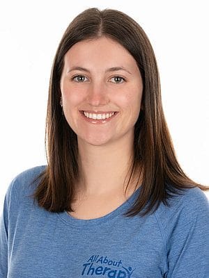 Smiling young woman in a blue All About Therapy t-shirt with long brown hair and natural makeup.
